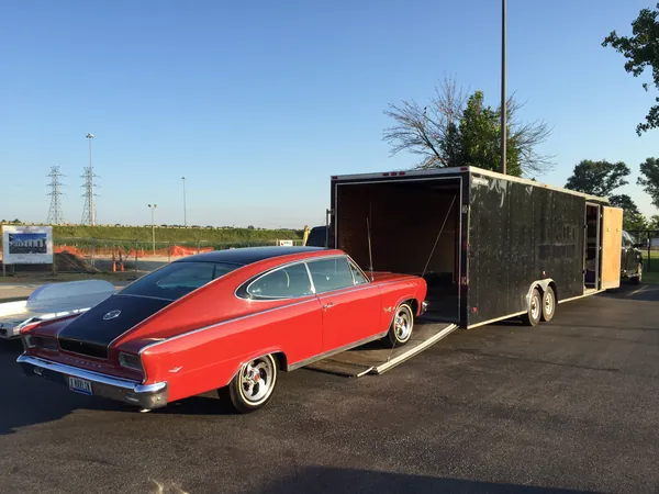 A red 1965 Rambler Marlin fastback parked beside a black single-car enclosed trailer with its ramp down